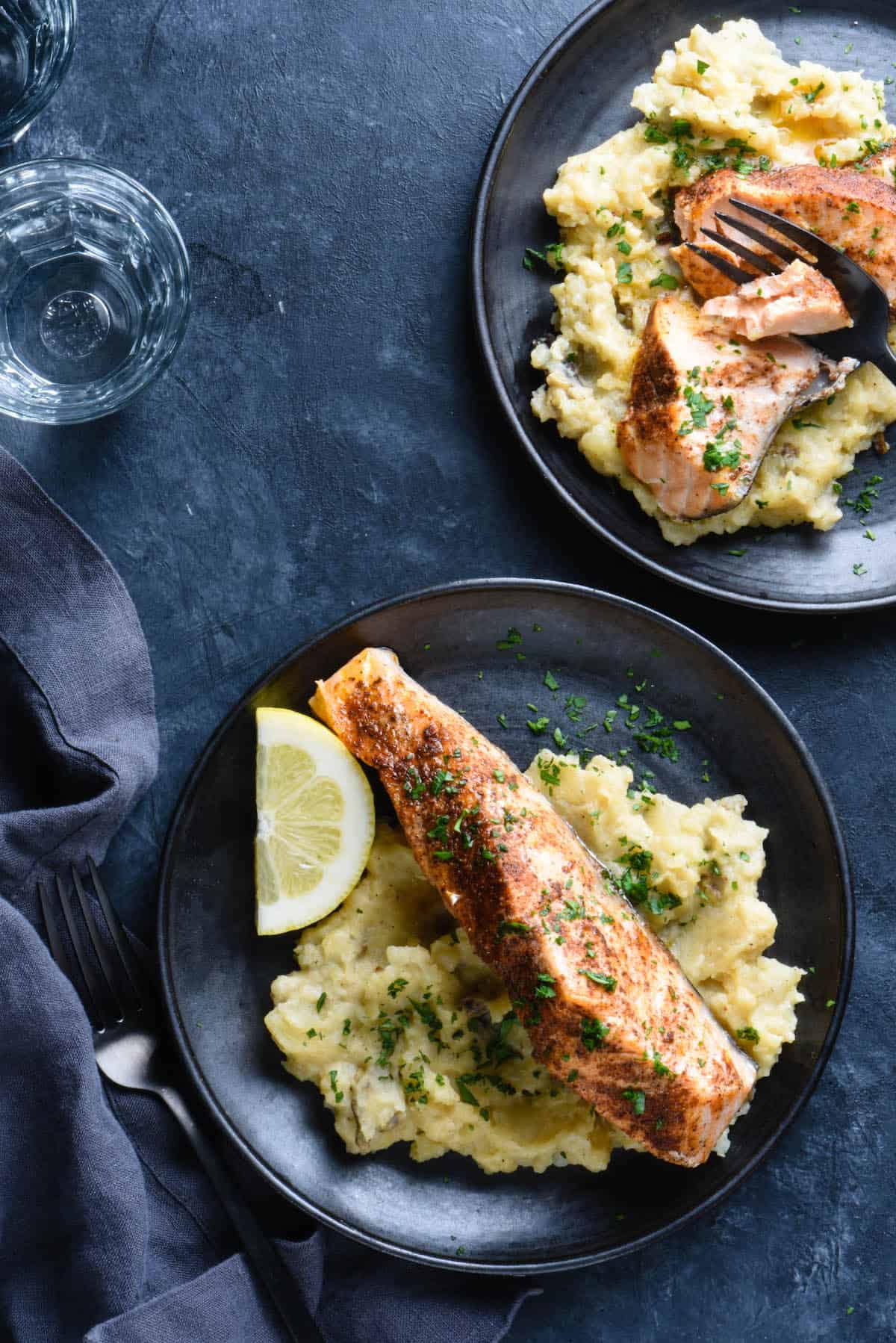 Dark background topped with two black plates. Plates are filled with instant pot salmon and mashed potatoes. Water glasses and napkin nearby.