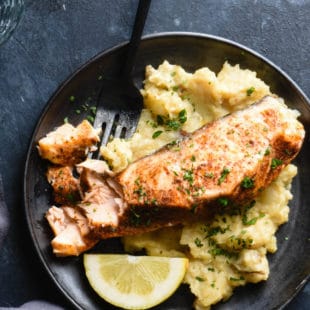 Black plate on dark background topped with mashed potatoes, salmon fillet and lemon wedge.