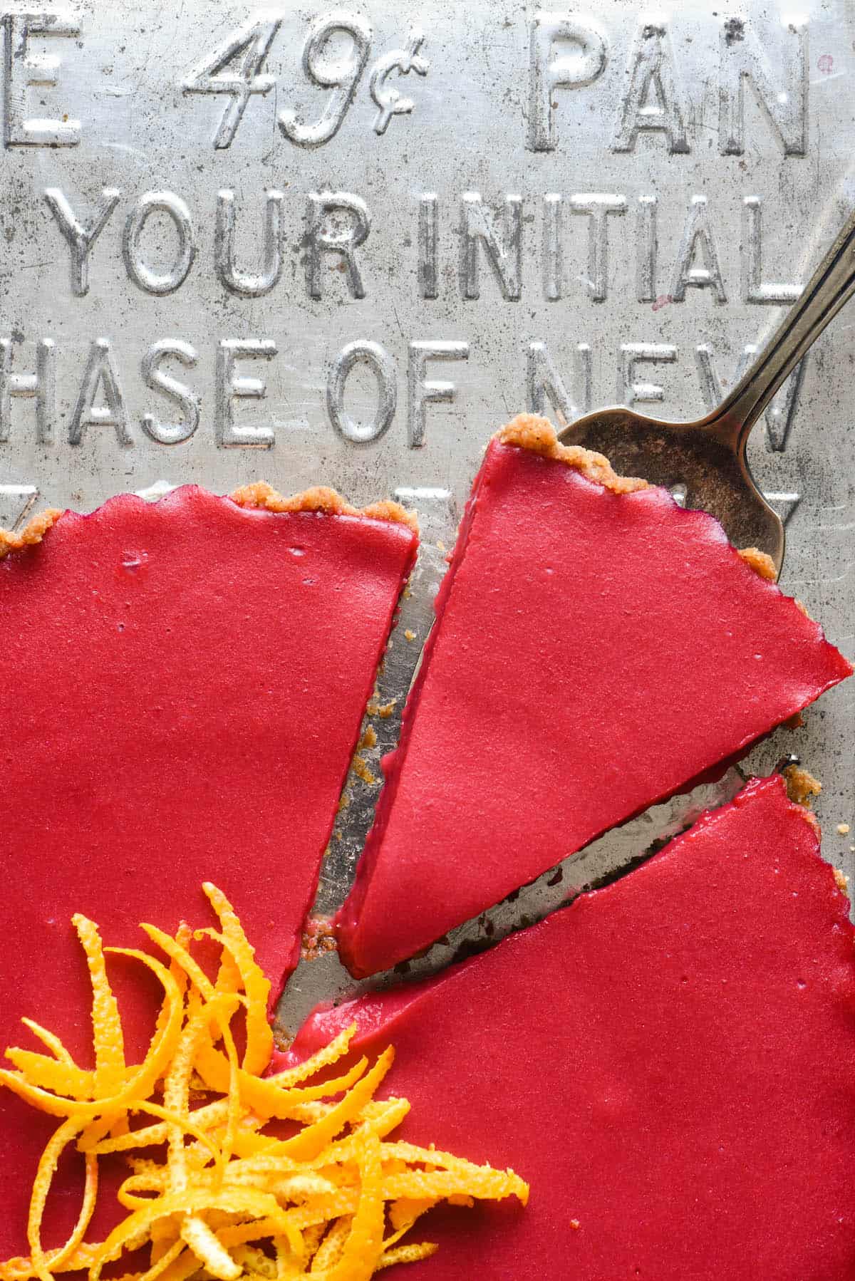 Closeup of one piece being cut out of a cranberry tart recipe, on a vintage baking pan. Orange peels garnish the tart.