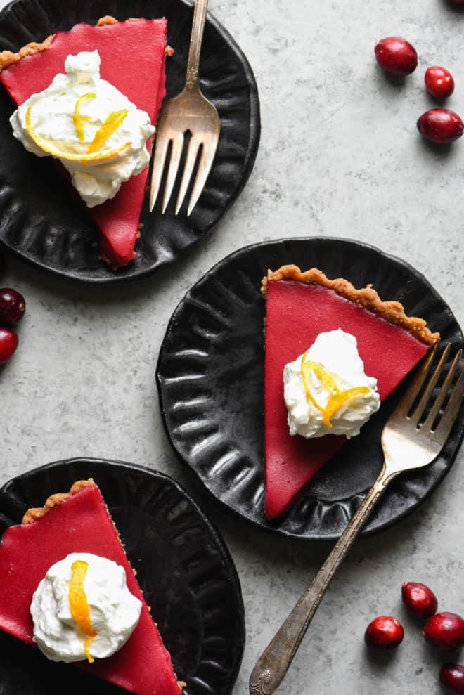 Overhead photo of three slices of cranberry curd on black plates, topped with whipped cream and orange zest.