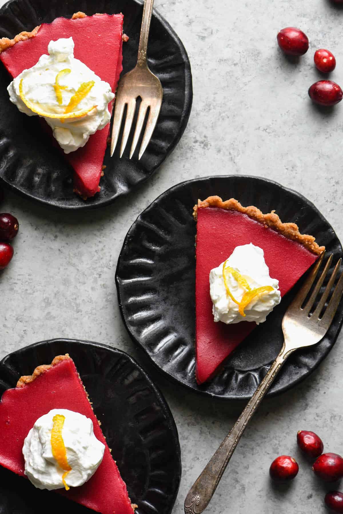 Overhead photo of three slices of cranberry curd tart on black plates, topped with whipped cream and orange zest.