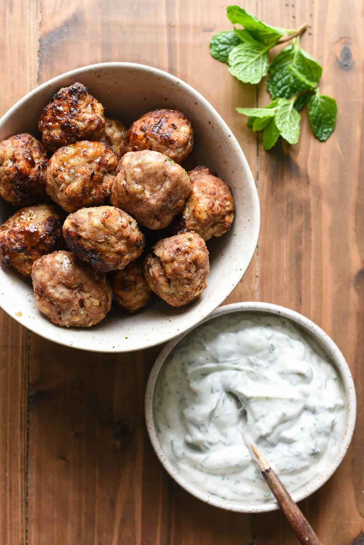 A bowl of air fryer turkey meatballs and a bowl of herbed yogurt sauce, with a mint sprig.