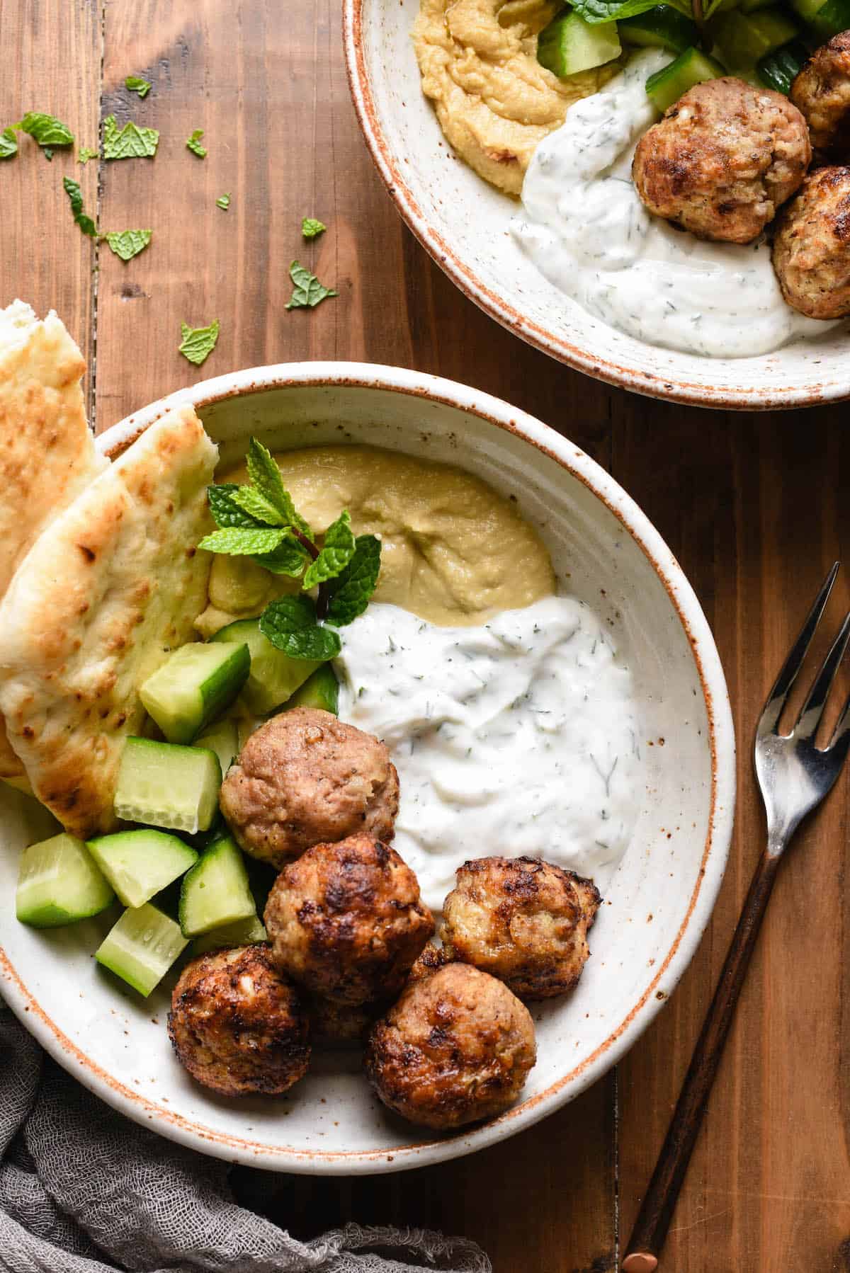 Two bowls filled with meatballs, diced cucumbers, herbed yogurt sauce, hummus and naan bread.