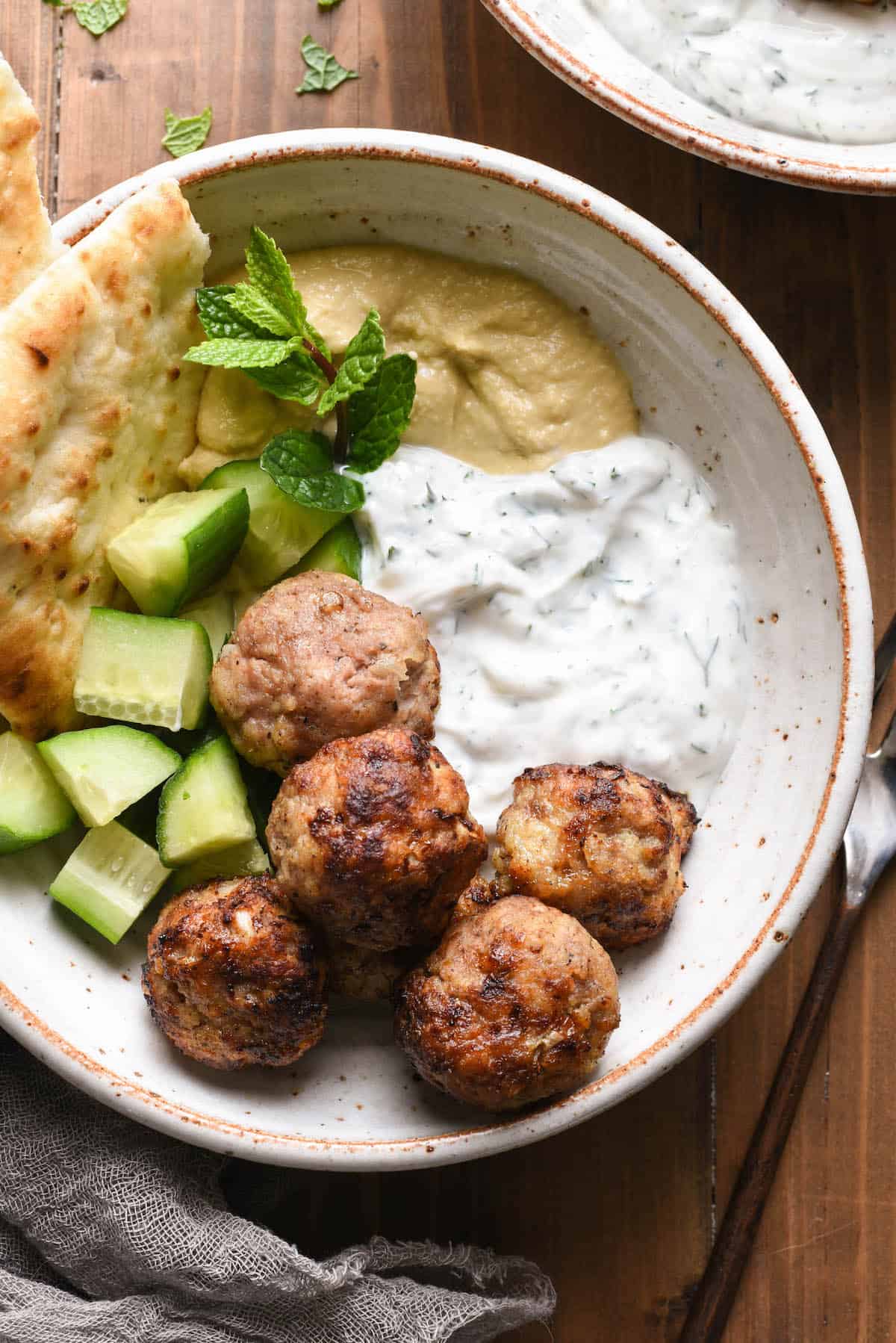 Overhead photo of rustic bowl filled with turkey meatballs, chopped cucumber, hummus, yogurt sauce, naan and a mint sprig garnish.
