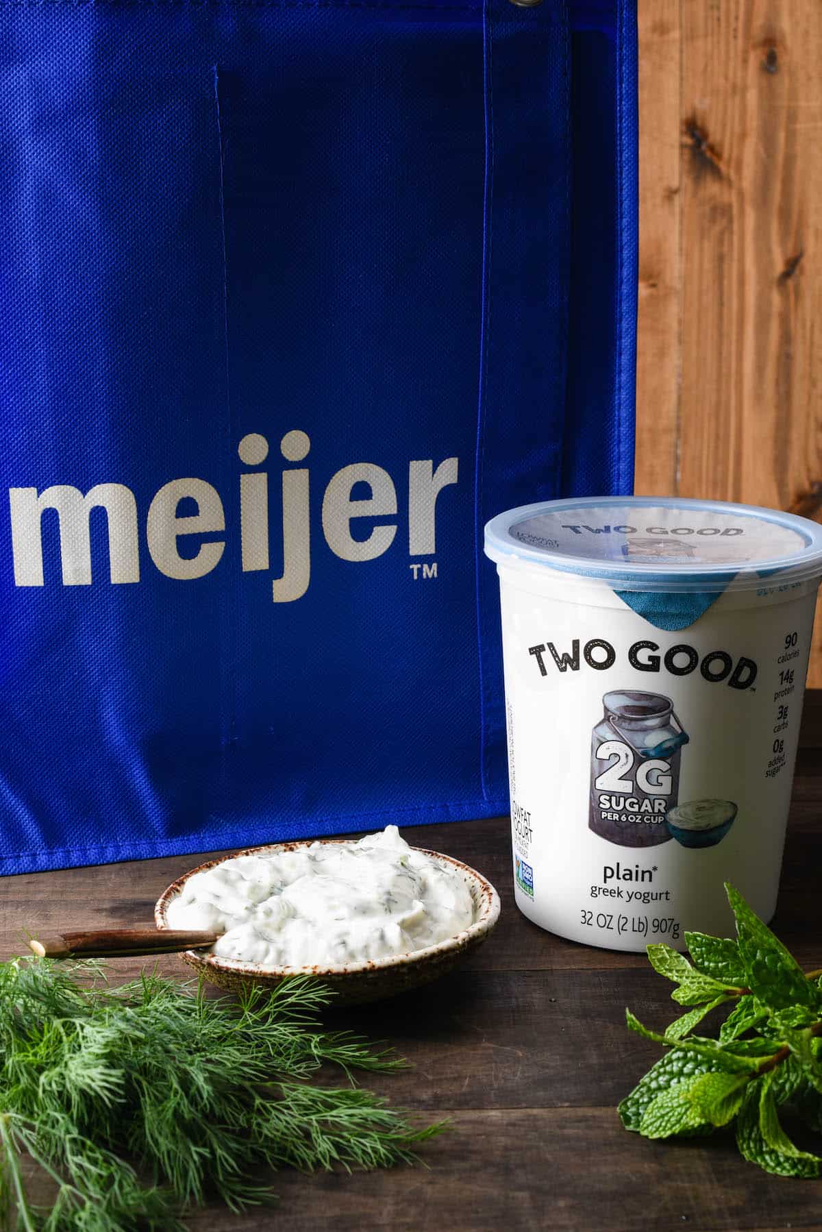 Container of Two Good Greek yogurt, bowl of yogurt sauce, mint and dill, with blue reusable Meijer bag in background.
