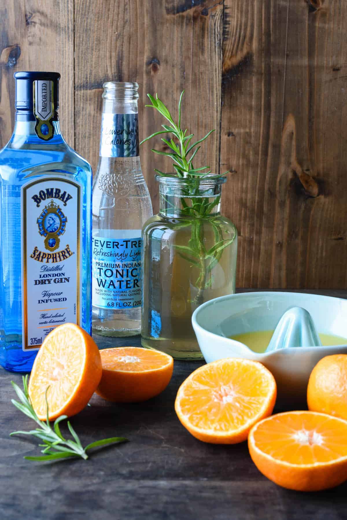 Gin and tonic water bottles, small bottle of rosemary simple syrup, ceramic juicer and cut oranges against wooden backdrop.