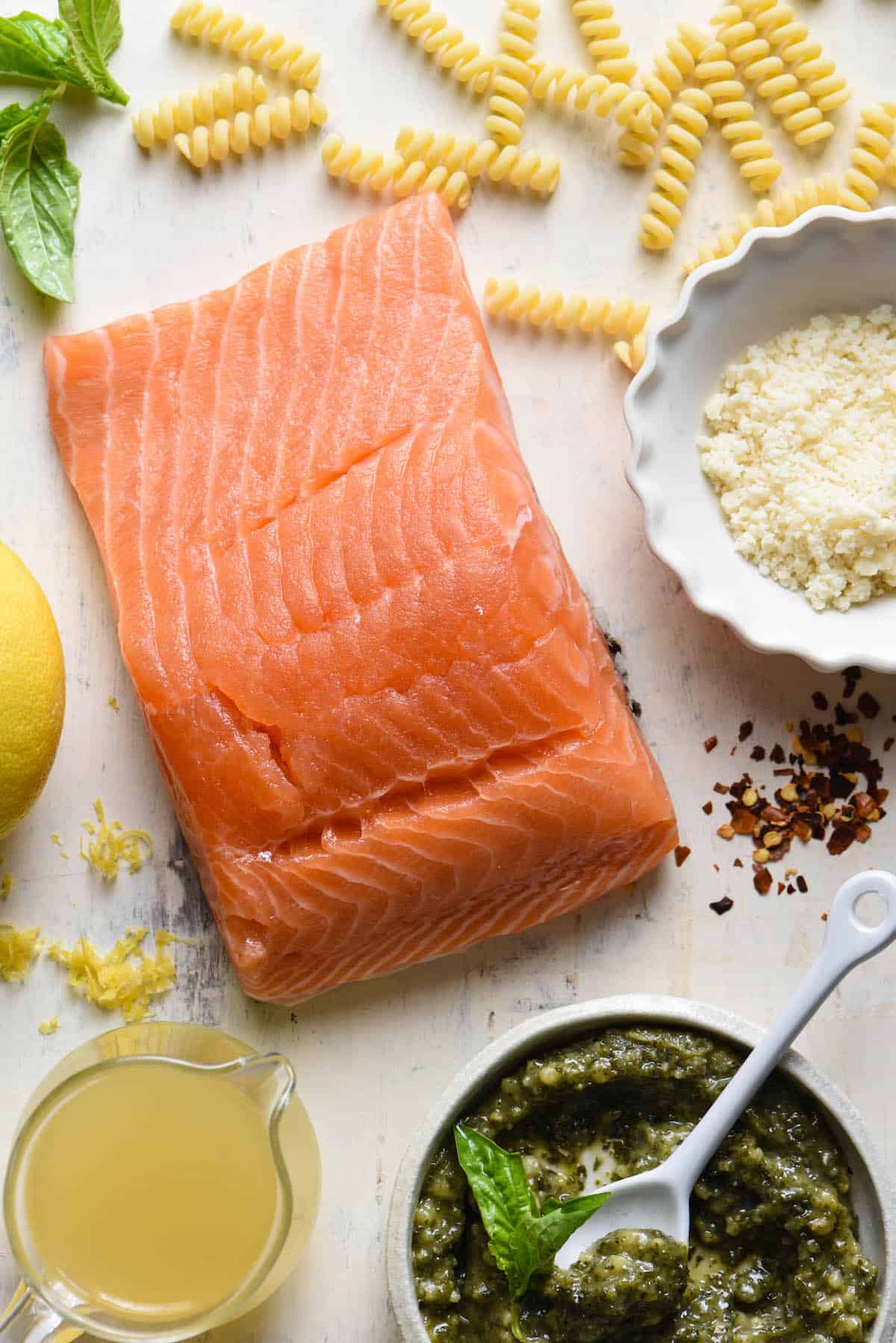 Overhead photo of ingredients for salmon pesto pasta, including a salmon fillet, dried pasta, basil leaves, Parmesan cheese, chicken broth, basil pesto, lemon zest, and red pepper flakes.