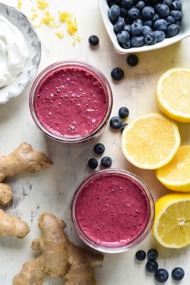 Overhead photo of two glasses of blueberry lemon smoothie alongside cut lemons, fresh blueberries, ginger root and yogurt.