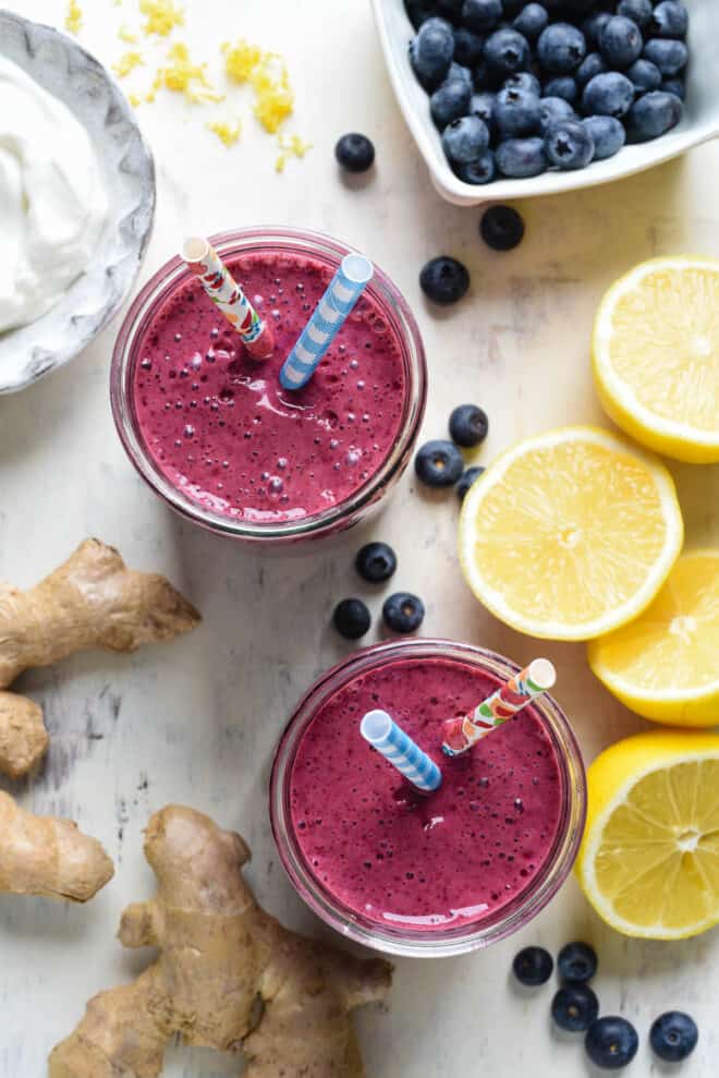 Overhead photo of two glasses of blueberry smoothie with yogurt, with fun straws, and fresh lemons, blueberries, yogurt and ginger root alongside.