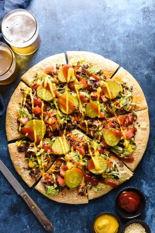 Overhead photo of a savory pie topped with meat and pickles, with two beers nearby.