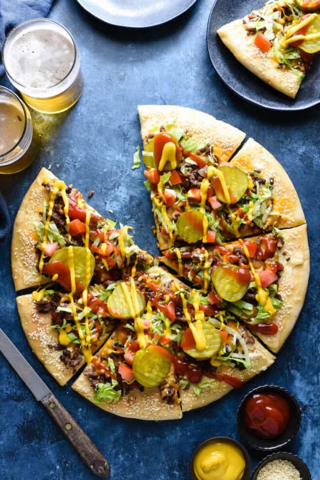 Overhead photo of cheeseburger pizza cut into eight slices, on blue background, with extra condiments nearby.