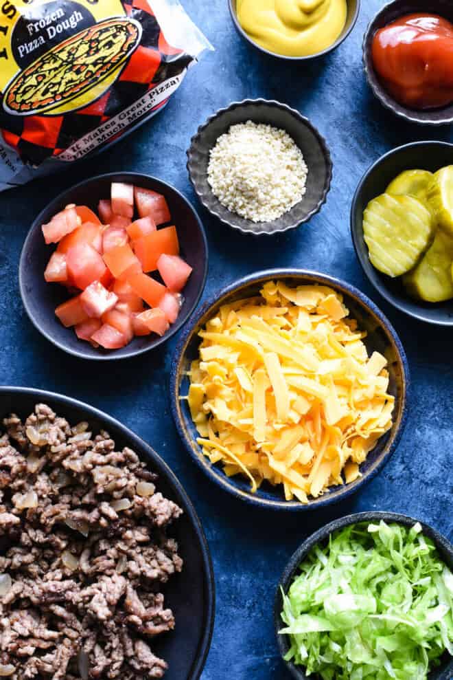 Blue tabletop with small bowls of ingredients like ground beef, shredded yellow cheese, diced tomatoes, pickles, sesame seeds, shredded lettuce, ketchup and mustard.