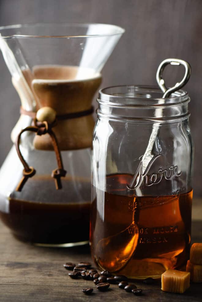 Quart sized mason jar half filled with brown syrup, with spoon in it. Chemex coffee maker is in background, caramel candies and coffee beans are in foreground.