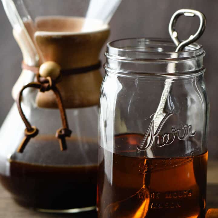 Quart sized mason jar half filled with brown syrup, with spoon in it. Chemex coffee maker is in background, caramel candies and coffee beans are in foreground.
