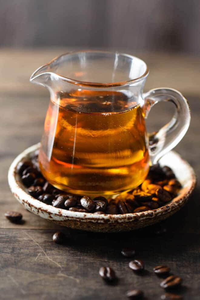 Small glass pitcher of brown liquid rests on plate filled with coffee beans.
