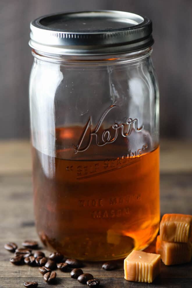 Quart sized mason jar half filled with brown syrup. Caramel candies and coffee beans are in foreground.