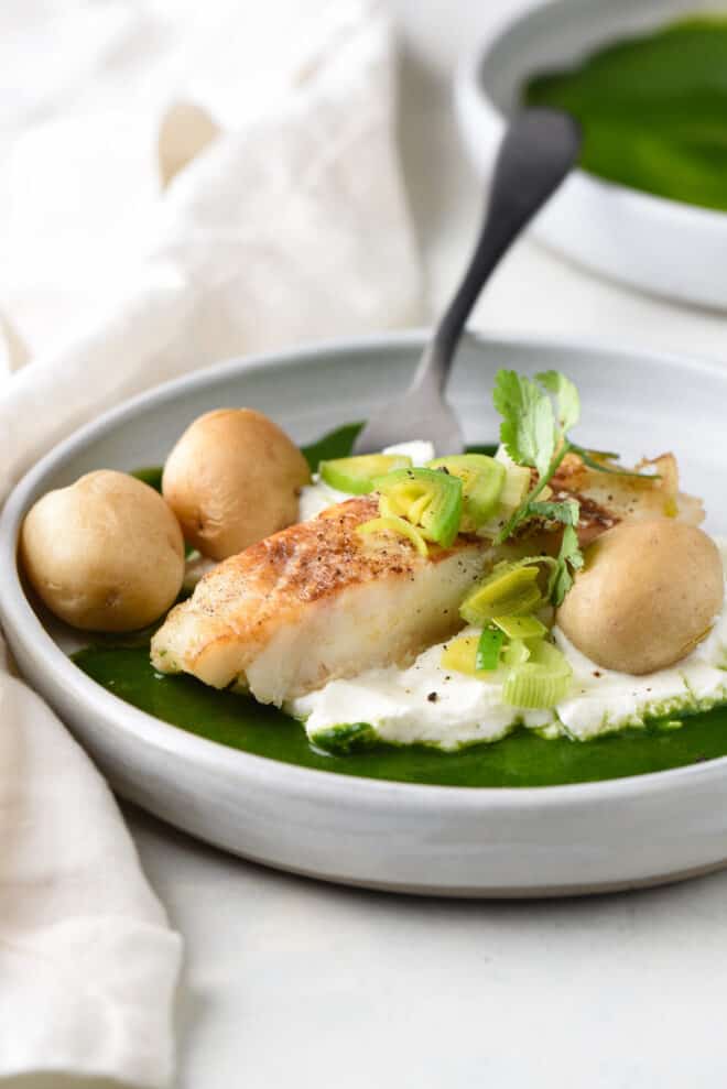 Shallow gray bowl filled with pan seared chilean sea bass, yogurt, a green sauce, and small boiled potatoes.