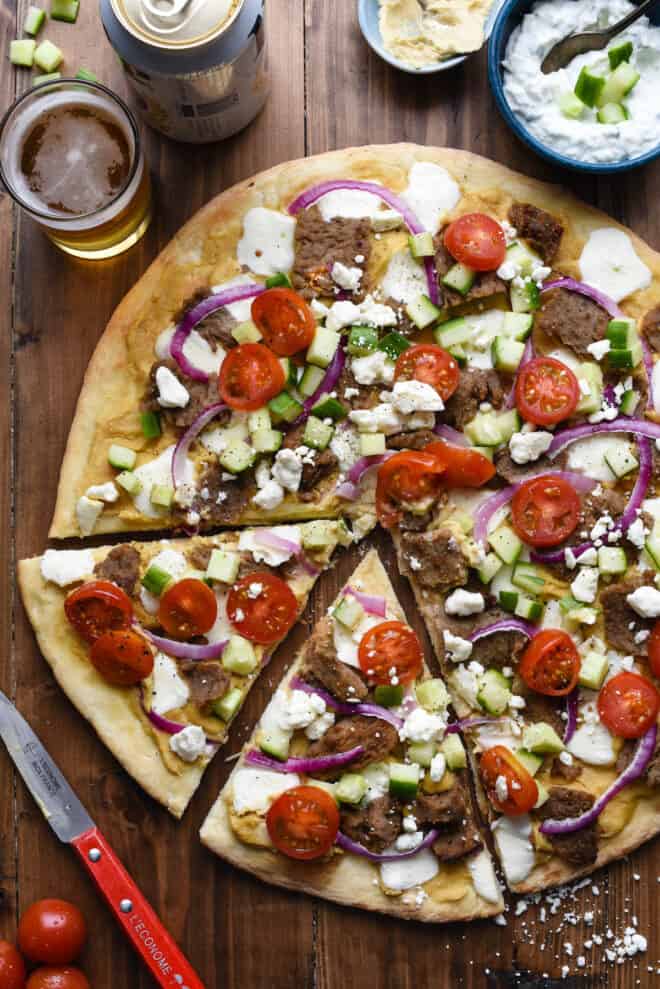 Overhead photo of gyro pizza with beer, yogurt sauce and hummus nearby.