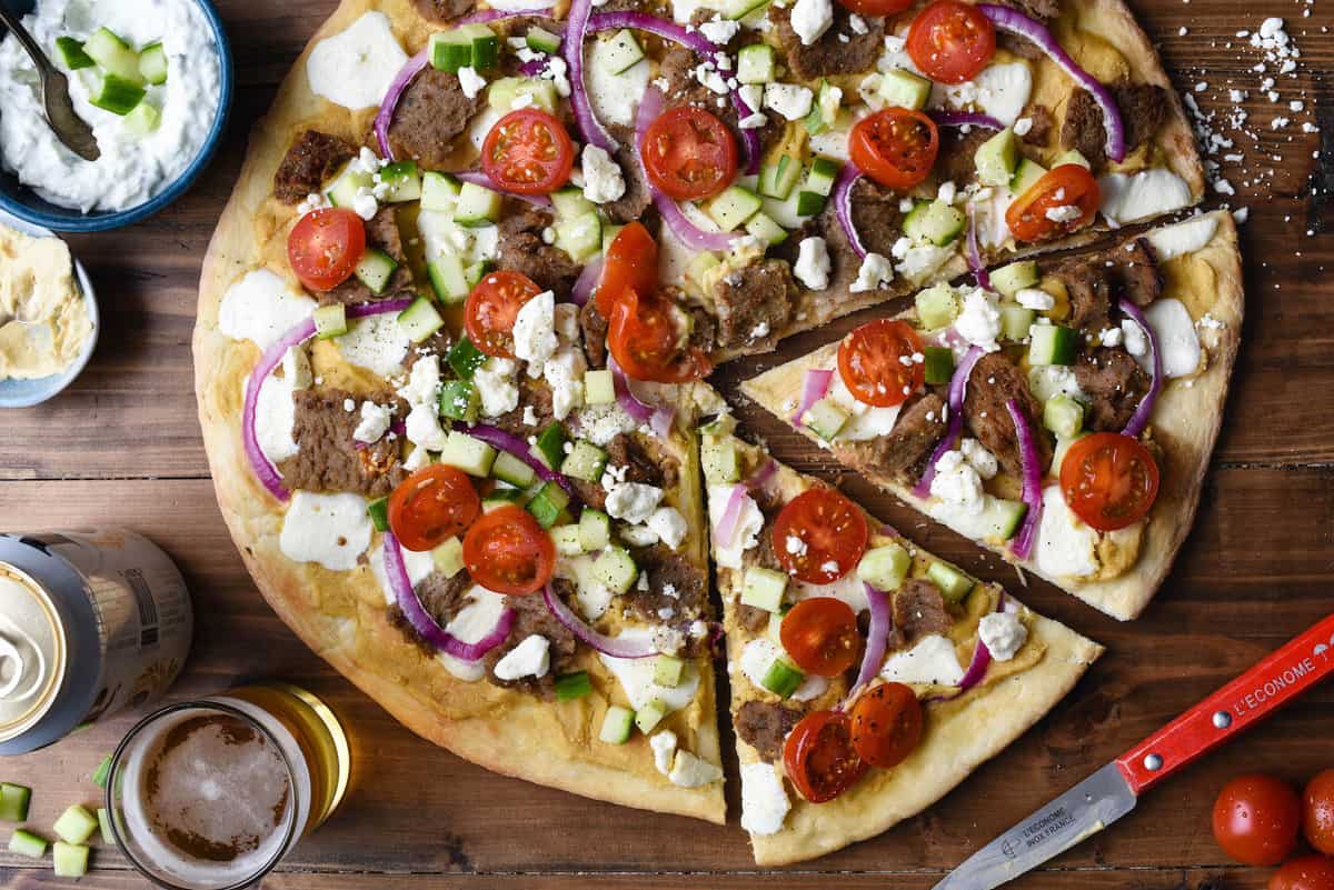Overhead photo of gyros pizza with beer, yogurt sauce and hummus nearby.