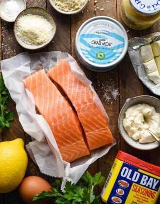 Overhead image of ingredients on a wooden surface, including salmon fillets, a can of crab meat, Old Bay seasoning, mayonnaise, cream cheese, lemon, Parmesan cheese, bread crumbs and salt.