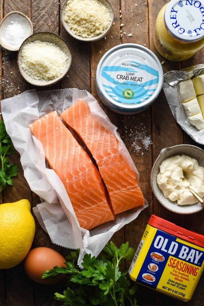Overhead image of ingredients on a wooden surface, including salmon fillets, a can of crab meat, Old Bay seasoning, mayonnaise, cream cheese, lemon, Parmesan cheese, bread crumbs and salt.