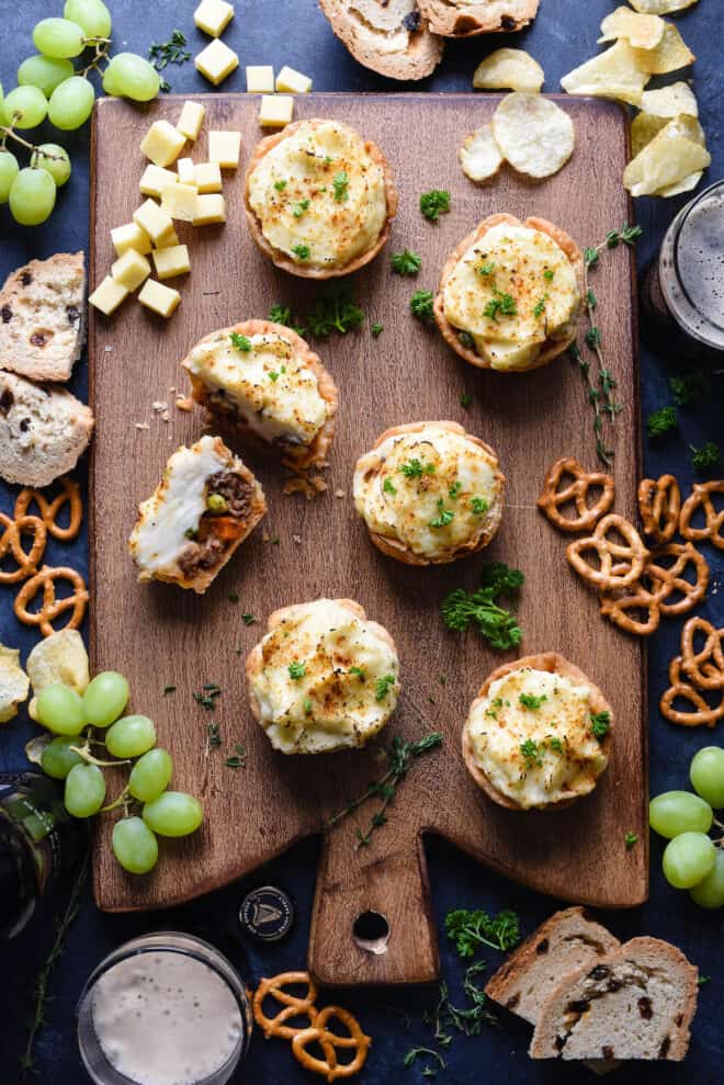 Small shepherd's pies on a wooden cutting board with party food like pretzels, chips, cheese and grapes.