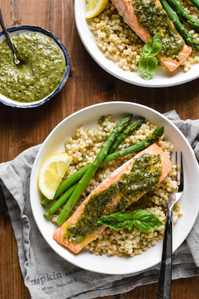 Shallow white bowls filled with pearl couscous, asparagus, and salmon topped with pesto sauce.