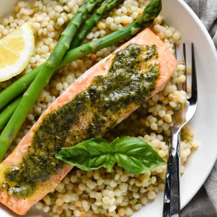 Shallow white bowl filled with pearl couscous, asparagus, and salmon topped with pesto sauce.