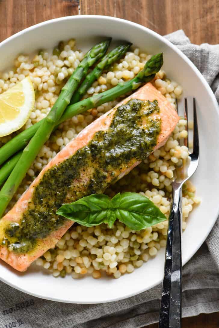 Shallow white bowl filled with pearl couscous, asparagus, and salmon topped with pesto sauce.