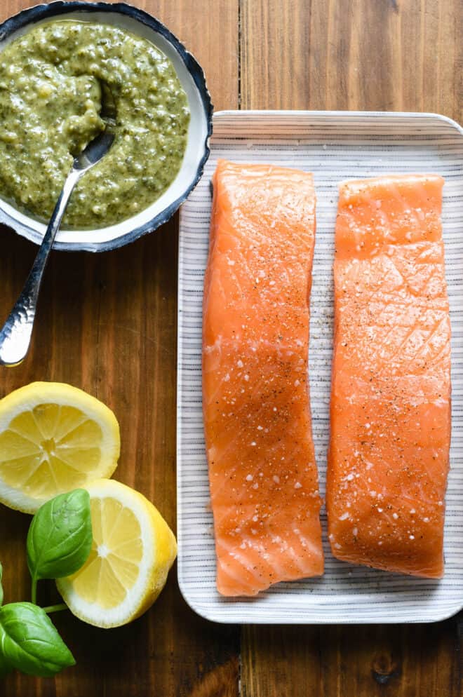 Small tray with two salmon fillets, bowl of basil pesto, halved lemon and fresh basil leaves, on wooden background.