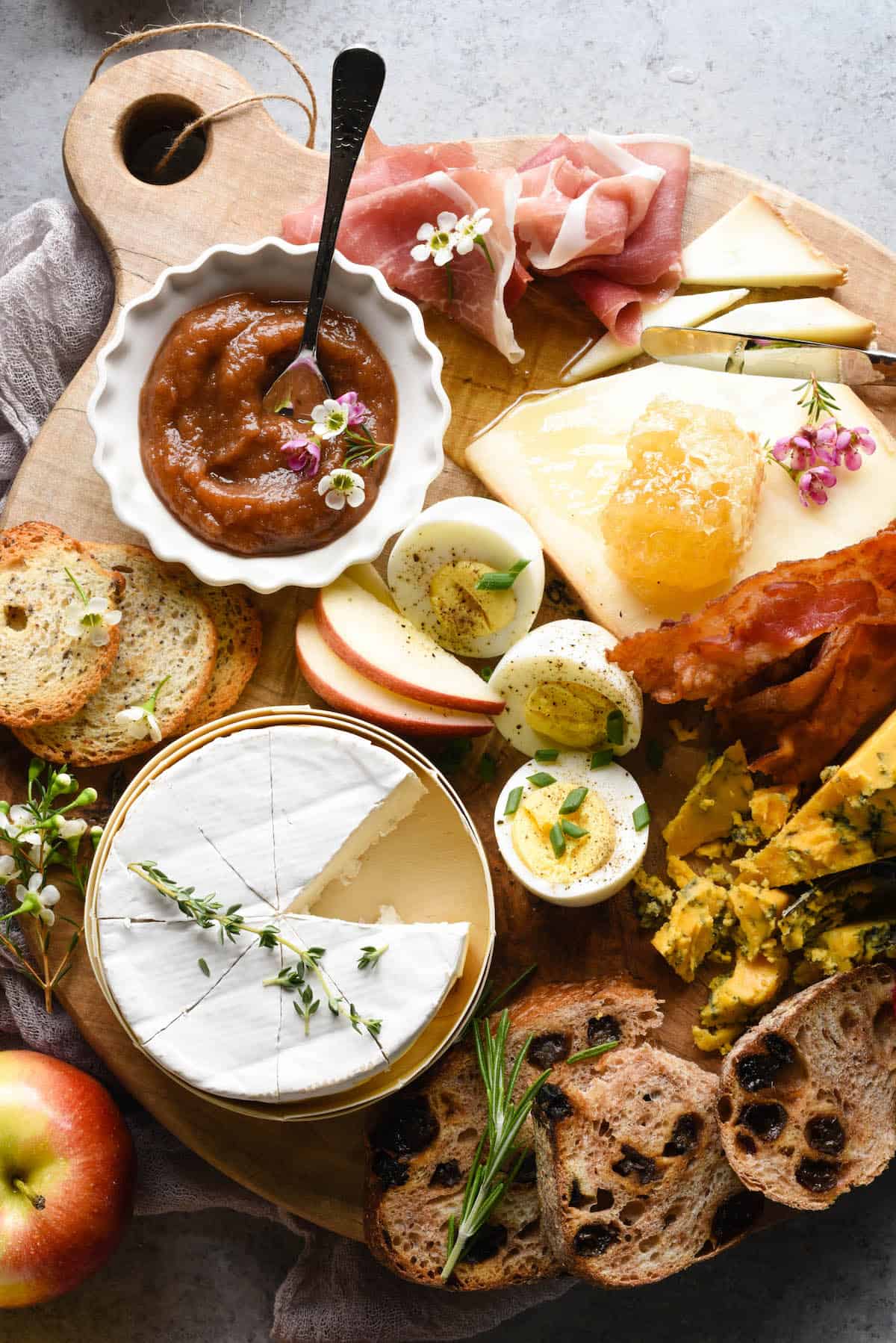 A breakfast charcuterie board arranged on a circular wooden board. Bread, cheese, hard boiled eggs, sliced apples, bacon and prosciutto cover the board.