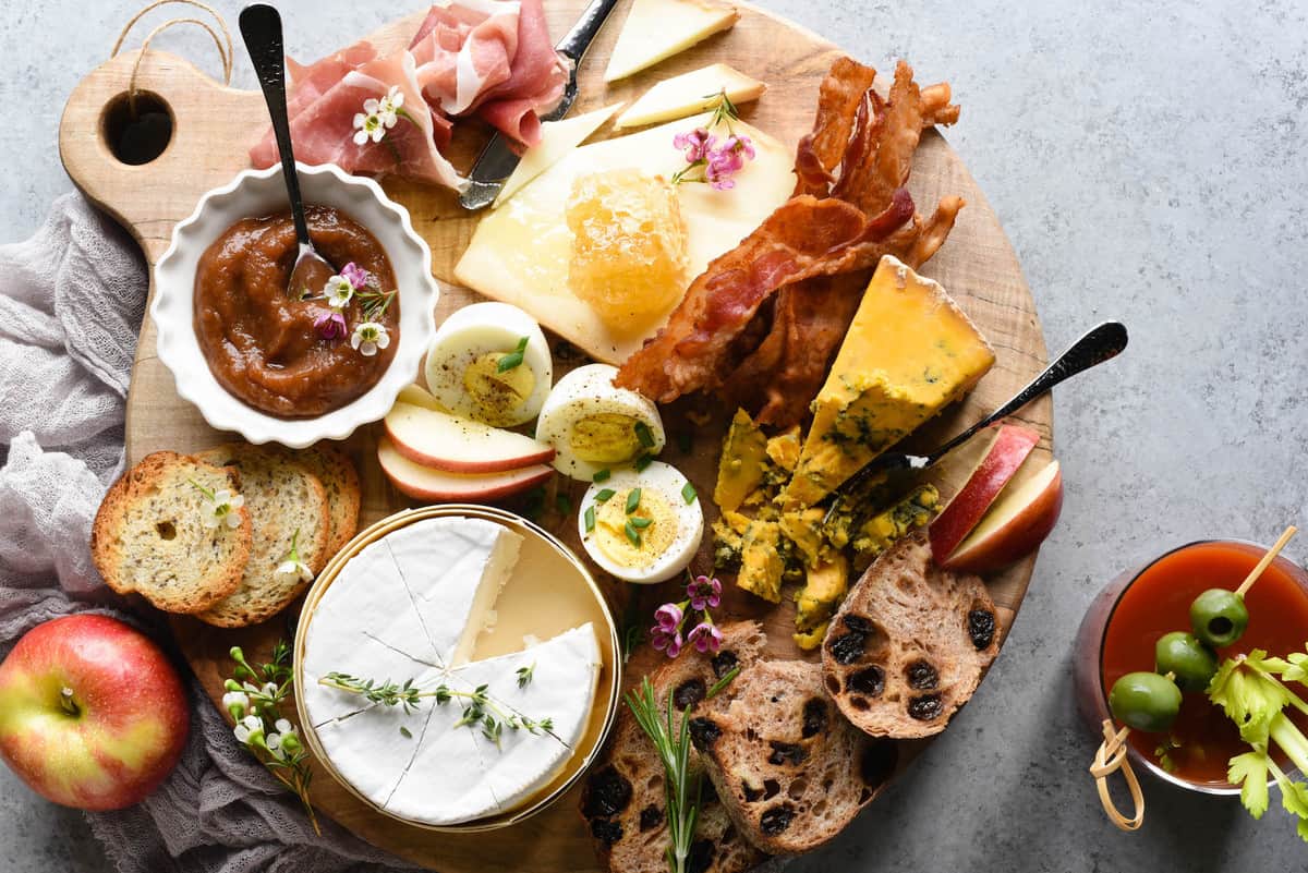 A brunch charcuterie board arranged on a circular wooden board. Bread, cheese, hard boiled eggs, sliced apples, bacon and prosciutto cover the board. A bloody mary is nearby.