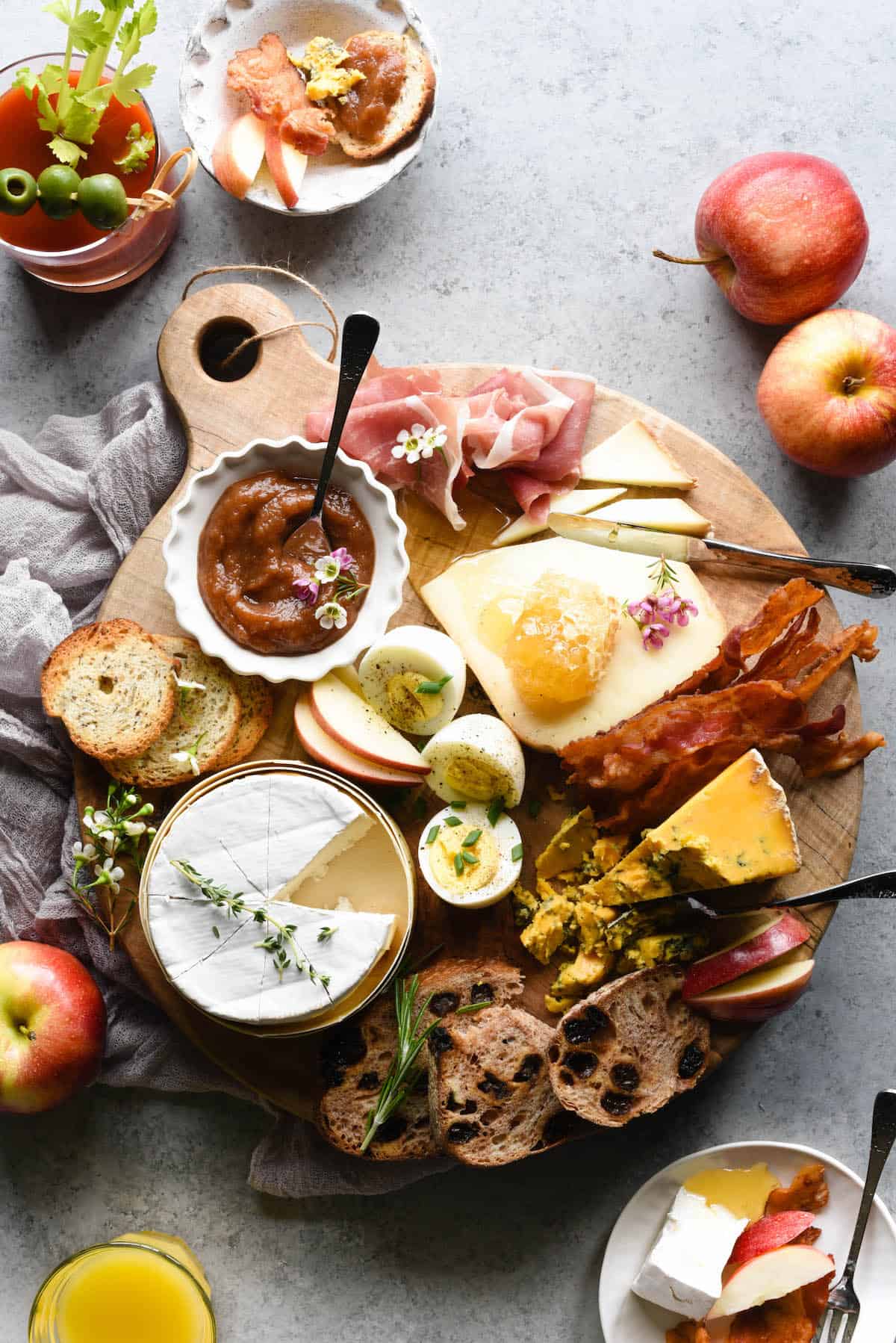 A breakfast charcuterie board - a round wooden cutting board filled with breakfast platter items like cheese, hard boiled eggs, bread, apple butter, bacon and prosciutto.