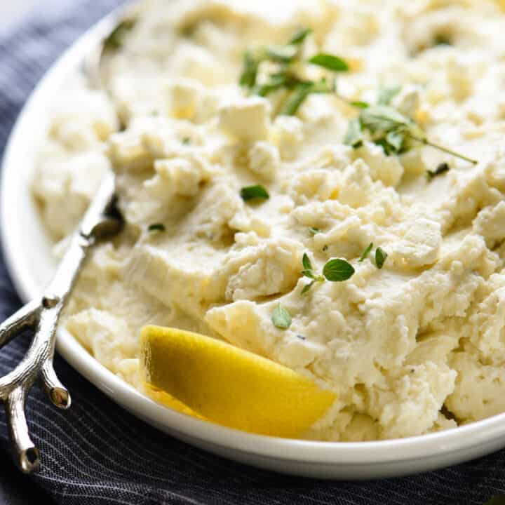 White oval platter of mashed potatoes garnished with herbs and lemon.