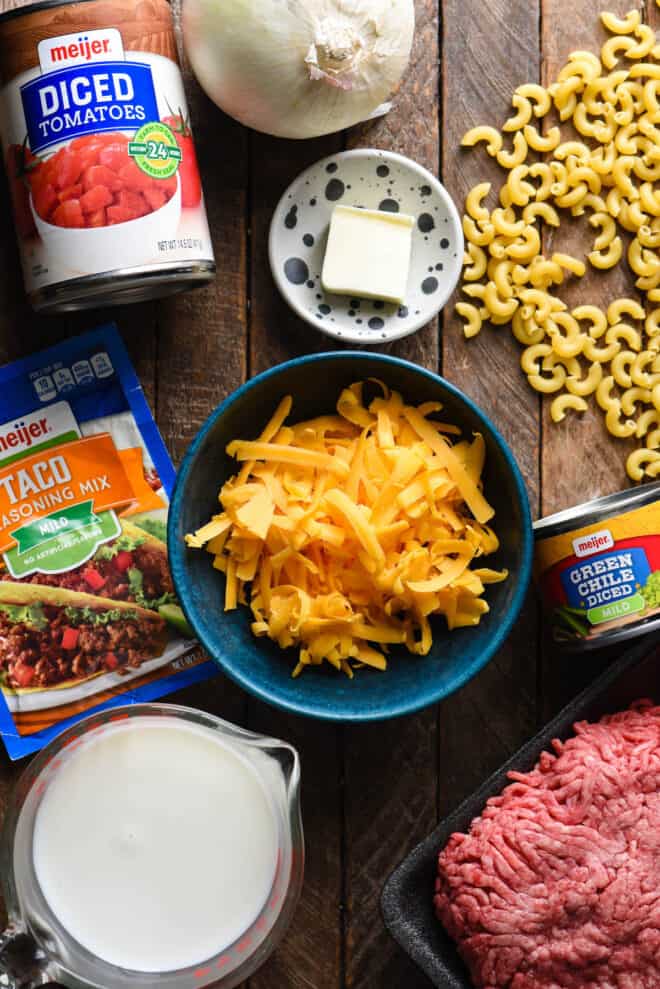 Wooden background with ingredients arranged on it, like shredded cheese, elbow noodles, taco seasoning mix, canned tomatoes and green chiles, butter, ground beef and onion.