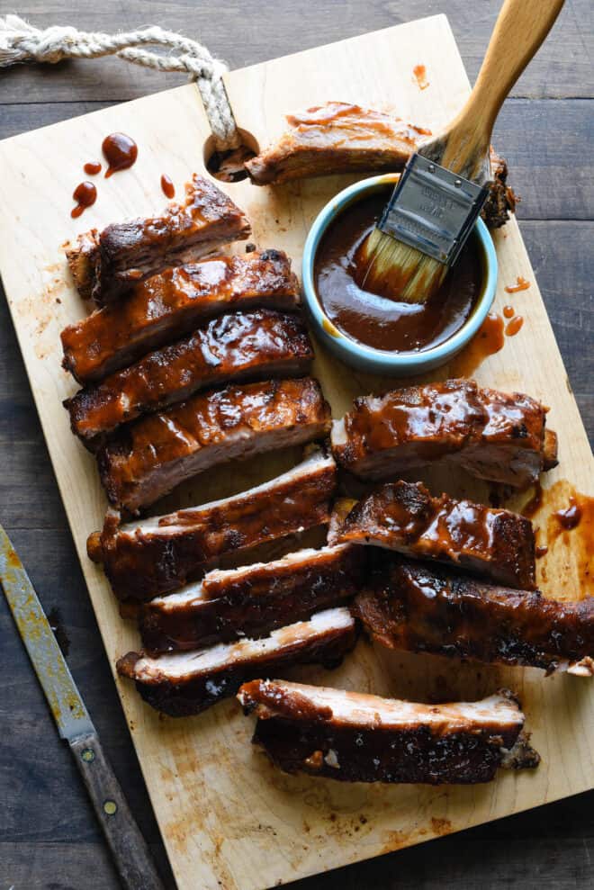 Wooden cutting board with cut racks of cooked baby back ribs glazed with barbecue sauce.