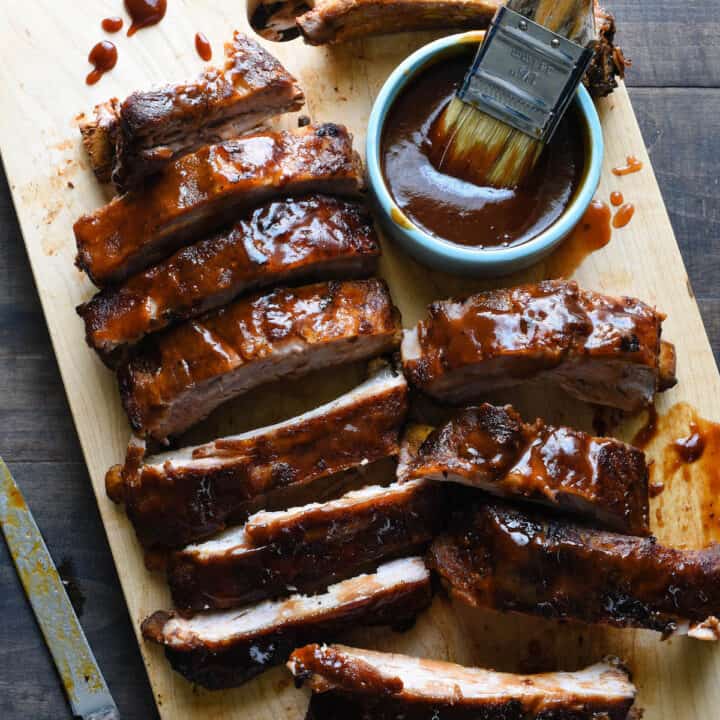 Wooden cutting board with cut racks of cooked baby back ribs glazed with barbecue sauce.