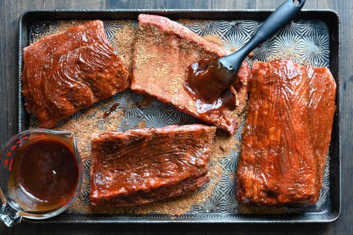 A textured baking pan with four raw sections of rib racks that have been rubbed with spices, being brushed with barbecue sauce.