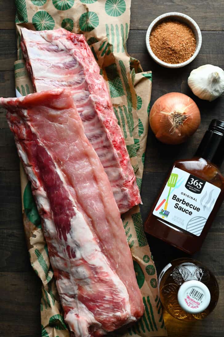 Ingredients for crock pot ribs laid out on a wooden surface, including two racks of ribs, onion, garlic, barbecue sauce and apple juice.