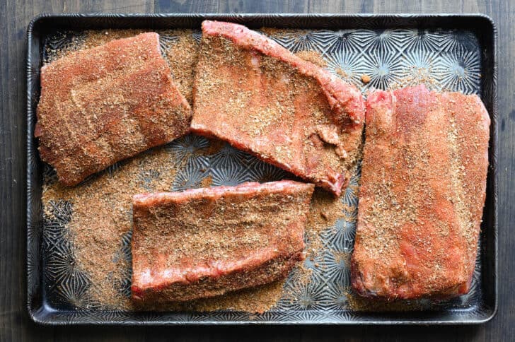 A textured baking pan with four raw sections of rib racks that have been rubbed with spices.