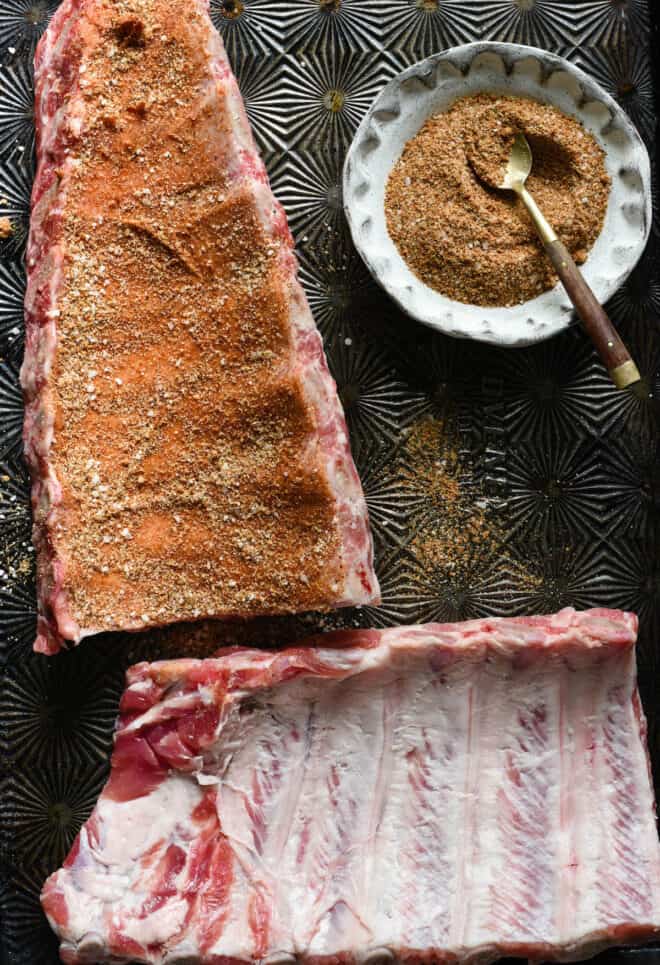 Textured baking pan with two racks of baby back ribs and small bowl of spice mixture. One rack of ribs is rubbed with spices.