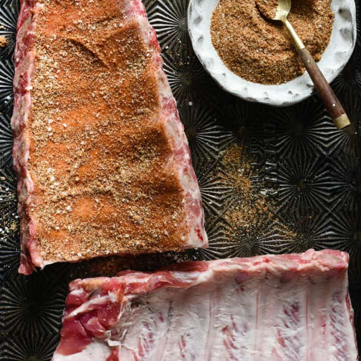 Textured baking pan with two racks of baby back ribs and small bowl of spice mixture. One rack of ribs is rubbed with spices.