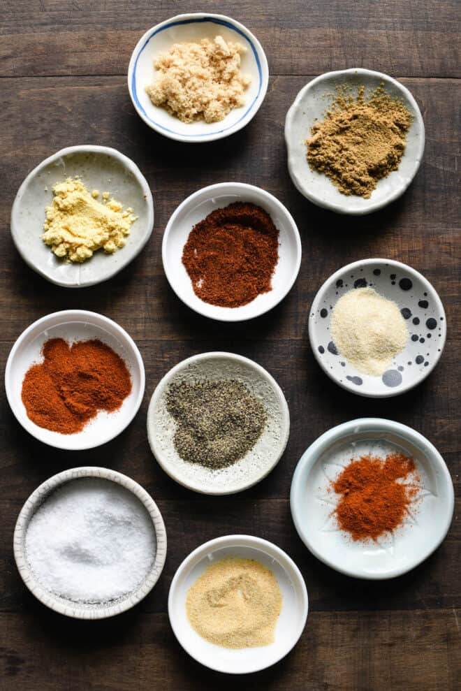 Ten small ramekins filled with various ground spices for rib rub, on wooden table.