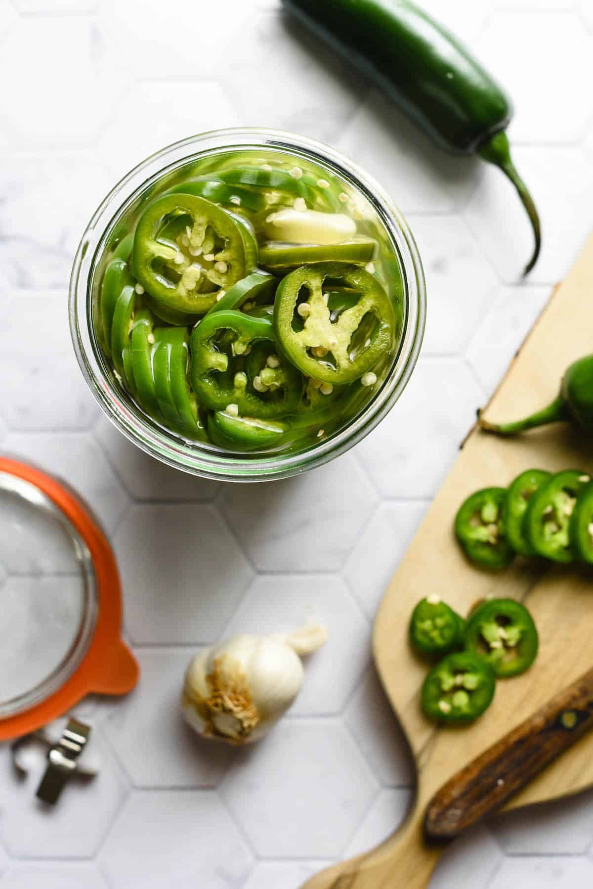 Jar of pickled jalapenos and small cutting board with knife against light background.