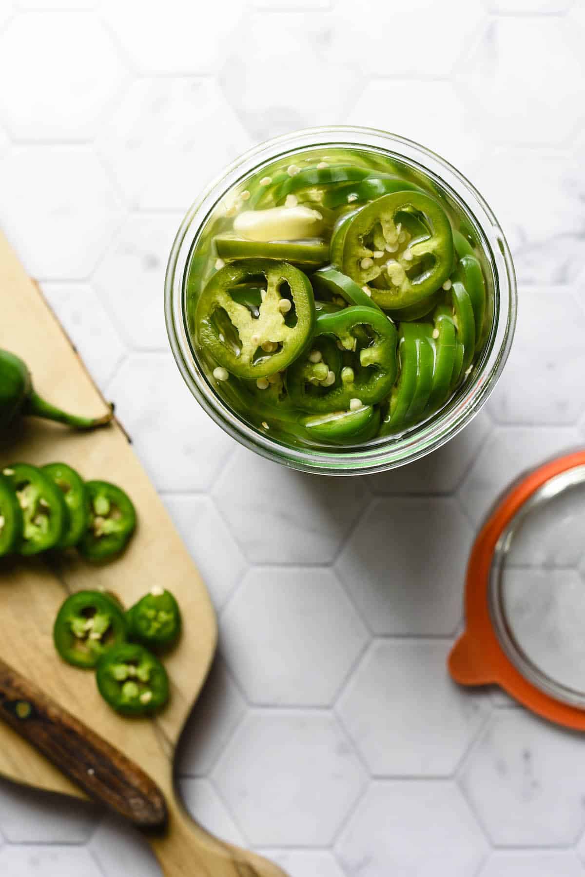 Jar of pickled jalapenos and small cutting board with knife against light background.