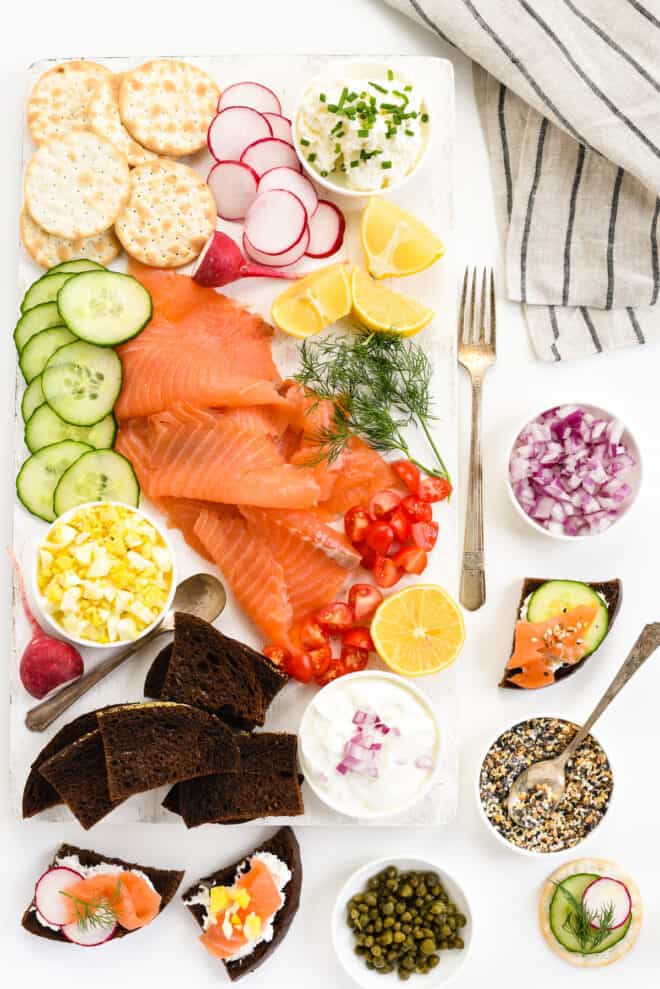 White table and cutting board topped with smoked salmon and garnishes like cucumber, capers, vegetables, cream cheese, bread and crackers.