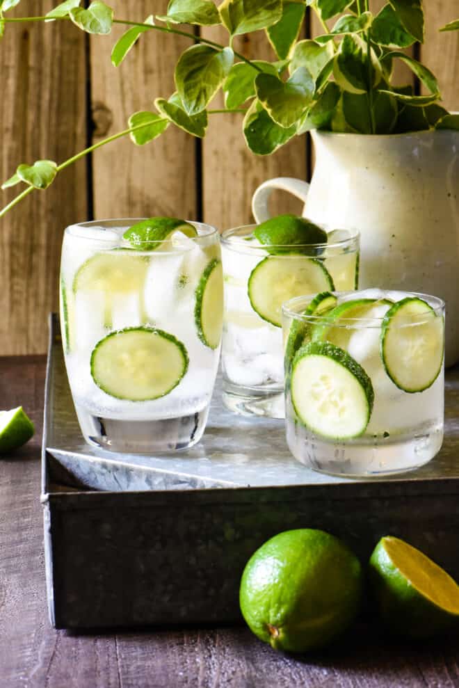 Three cocktails on a tray, garnished with lime wedges and cucumber slices.