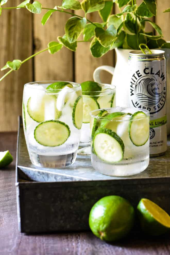 Three glasses filled with limes, cucumbers and clear liquid, with a can of White Claw next to them.