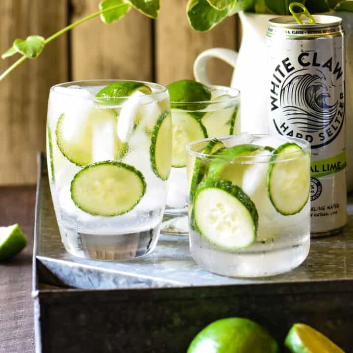 Three glasses filled with limes, cucumbers and clear liquid, with a can of White Claw next to them.
