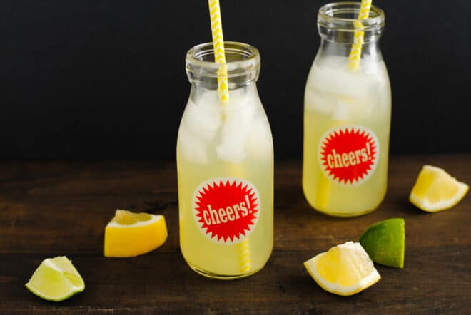 Two small glass bottles with red "cheers" stickers on them, filled with icy lemonade and yellow straws.